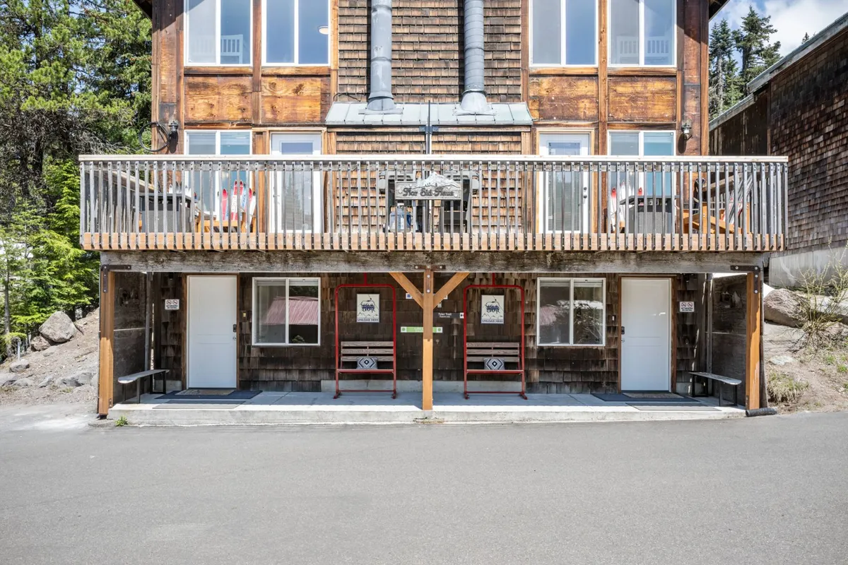 The exterior of a rustic yet welcoming lodge featuring a wooden balcony, large windows, and a prominent entrance sign. The inviting balcony suggests a social, communal atmosphere with easy transition between indoor comfort and outdoor adventure.