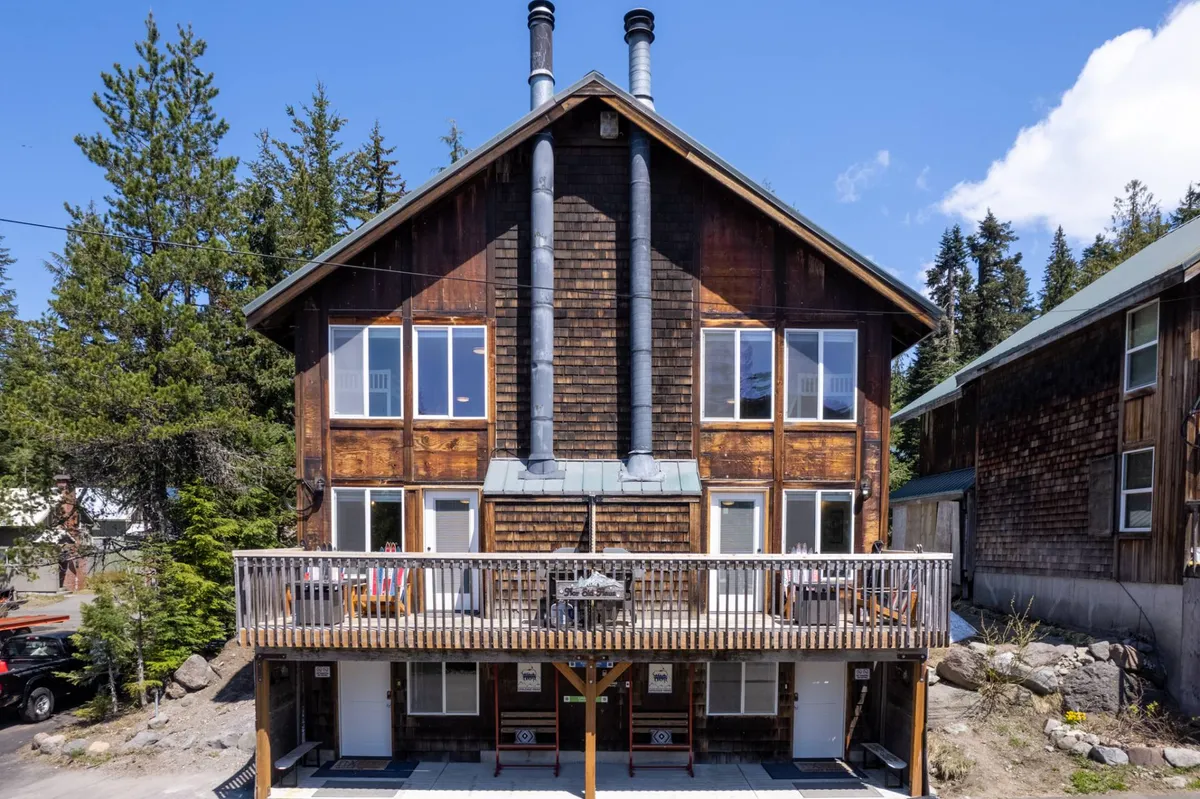 The image showcases the exterior of a spacious mountain town house with inviting wooden balconies, multiple chimneys, large windows, and rustic wood siding. The scene evokes a cozy, welcoming atmosphere, perfect for gatherings and enjoying the natural surroundings. The balcony offers sweeping sunset views, ideal for relaxing or savoring a morning coffee while immersed in the forested mountain environment.