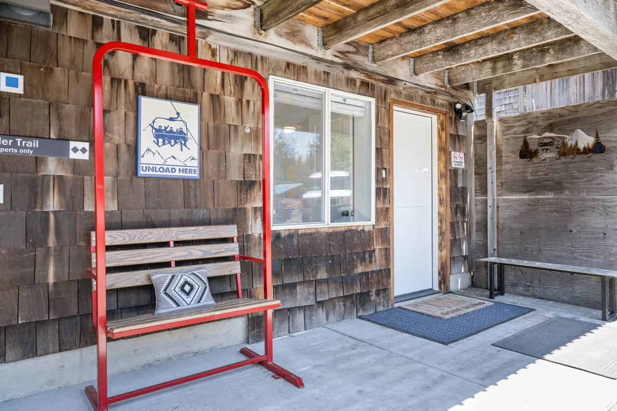 An exterior shot showcasing the front door area with a red unloading sign, a wooden bench, decorative wall art, and the entrance to the property. The scene conveys a sense of arrival and the practical setup for guests with gear or pets.