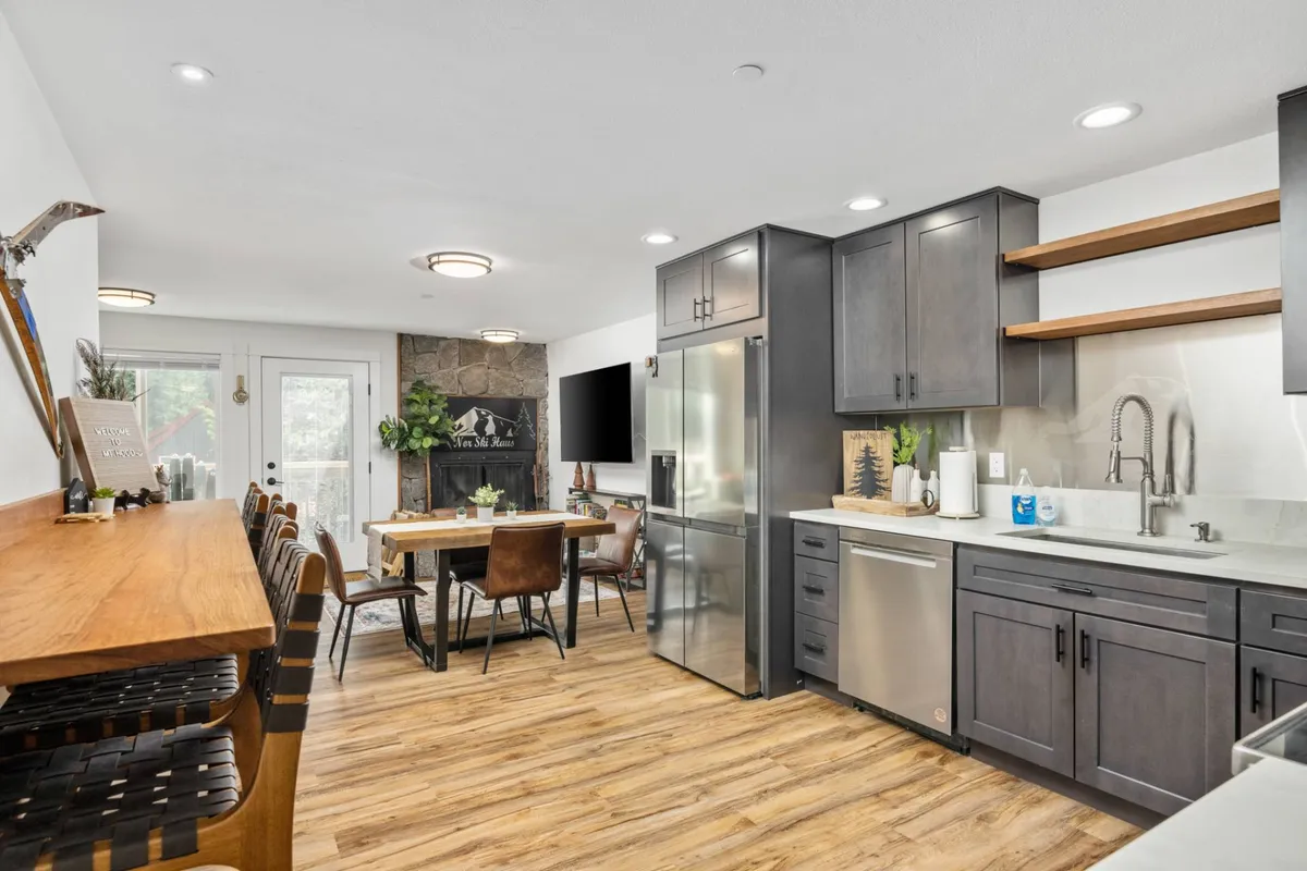 A spacious, open-plan interior showcasing a kitchen island, dining table, and cozy fireplace. Large windows flood the room with light, and the setup invites both cooking and socializing in a warm, communal atmosphere. The seating arrangement encourages conversation around the dining table, while the fireplace provides a focal point for relaxation.