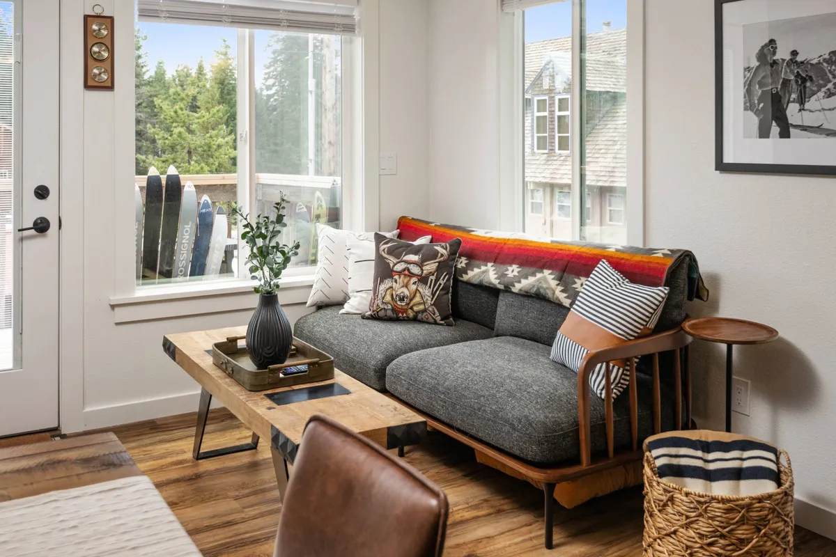A cozy living area with a plush sofa, coffee table, large windows, and a leafy plant. The windows provide ample natural light and a sense of connection to the outdoors, reinforcing the notion of transitioning from outside in—a subtle nod to arriving, parking, and settling in.