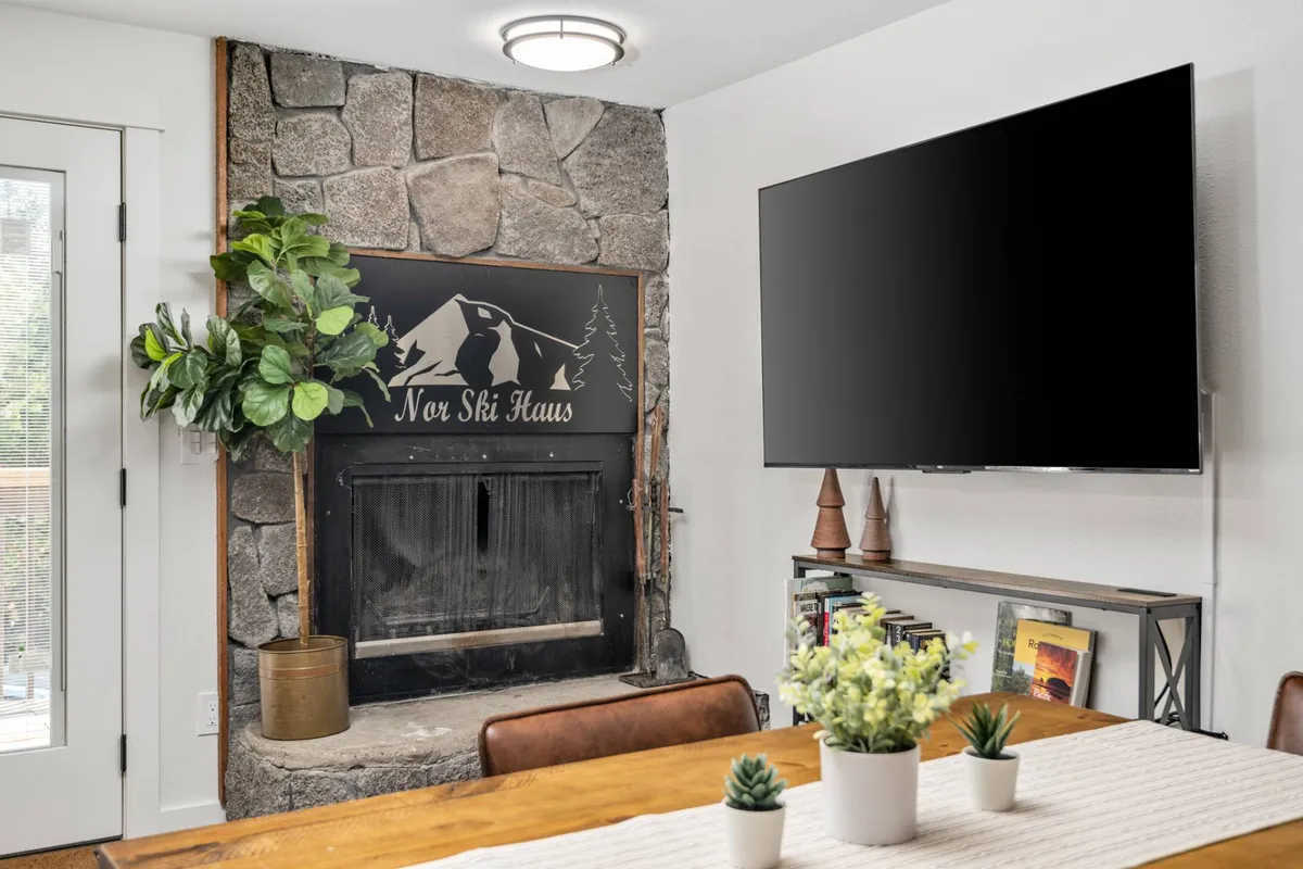 A warm, welcoming interior centered around a stone fireplace, expansive dining table, and a large TV. Decorative plants and soft lighting create a cozy ambiance, ideal for game nights, movie marathons, or casual dining with your group.