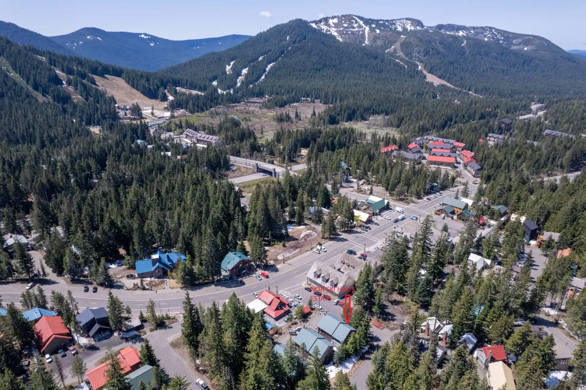 A vivid vista capturing the heart of Government Camp: a cluster of charming buildings flanked by intersecting roads, with the dramatic backdrop of a mountain range. Visible are hints of local shops, trails, and areas for outdoor activities, suggesting walkability and easy access to both nature and village life.