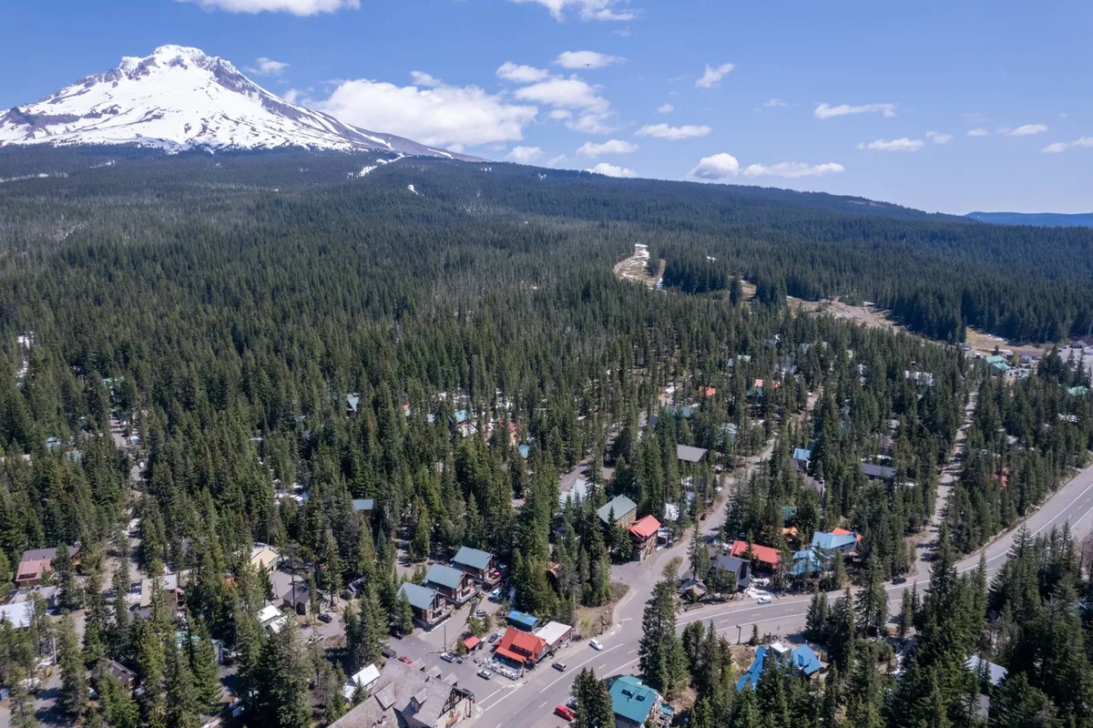 A sweeping view of a snow-capped mountain (Mt. Hood) framed by dense evergreen forest, with colorful chalet-style houses nestled below and a winding road leading toward the peaks. The image exudes tranquility and adventure, highlighting both the natural landscape and the proximity to adventure.
