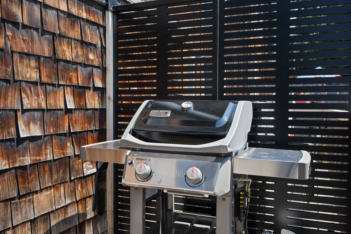 An exterior deck area with a modern grill set against a wooden wall and screening panels. The setup is ready for barbecuing and entertaining, featuring plenty of space for outdoor gatherings and summer celebrations.