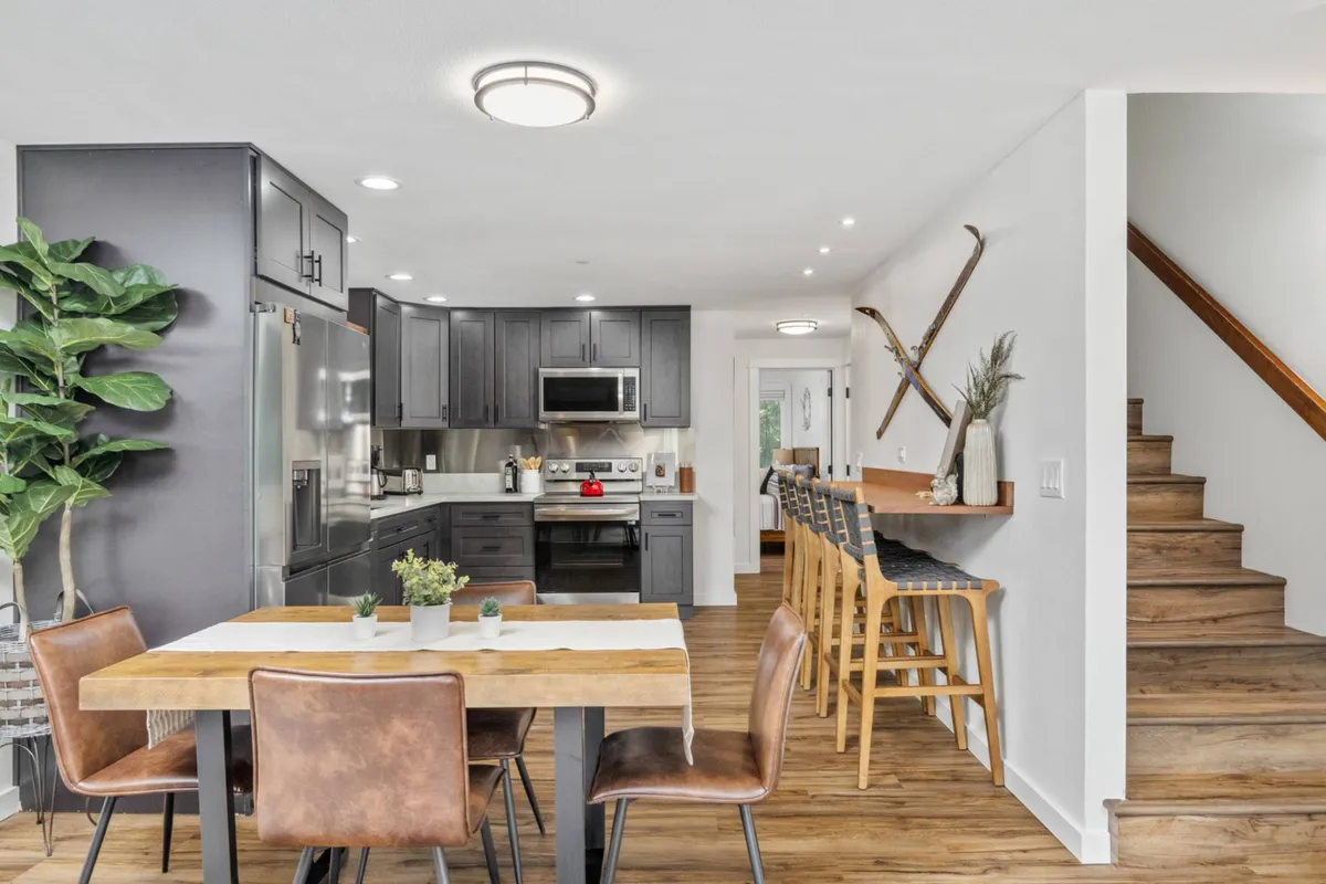 The focus here is on a large dining table and kitchen island, surrounded by an inviting, cozy atmosphere. The view includes the staircase, reinforcing the sense of a spacious, multi-level home. The scene exudes warmth and is clearly meant for group meals, family game nights, and social gatherings, with plenty of seating and surfaces for shared activities.