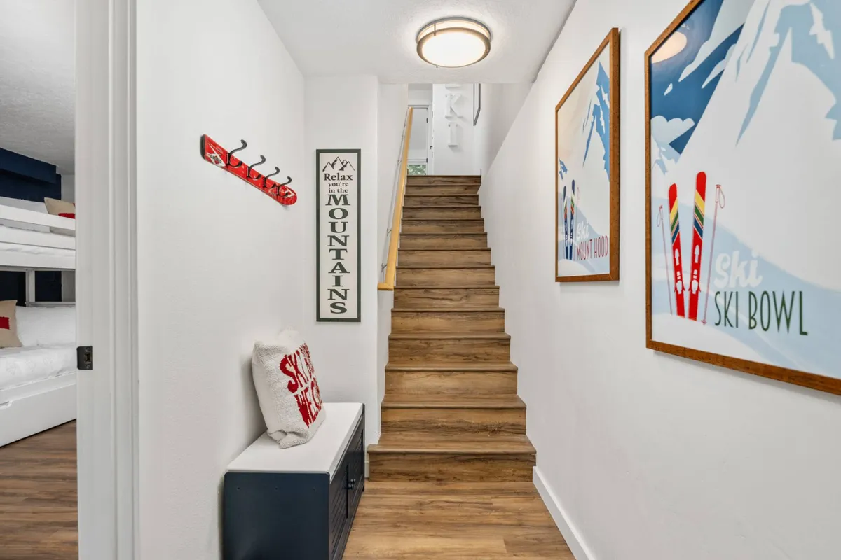 An interior hallway scene featuring a staircase, a bench with throw pillows, wall hooks for outerwear or gear, and wall art. This area serves as a transitional space for guests to prepare for or return from outdoor activities, reinforcing the mudroom and gear storage narrative.