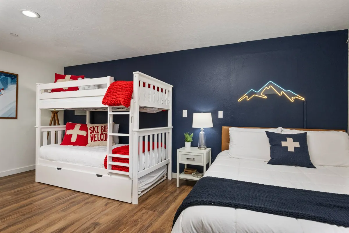 A playful and inviting family bedroom with a bunk bed and a double bed, both adorned with red throw blankets and mountain-themed artwork. The neon mountain light adds a whimsical touch, while the setup is clearly designed for family-friendly stays or group sleepovers.