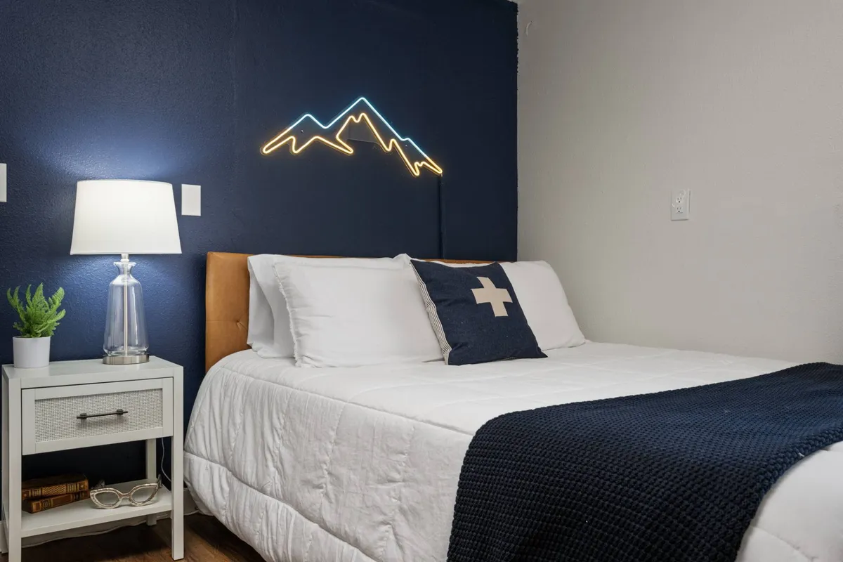 A tranquil bedroom scene featuring a bed with crisp white linens, a minimalist nightstand with a lamp, and a glowing neon mountain light above the headboard. The room radiates calm and serenity, perfect for restful nights after mountain adventures.